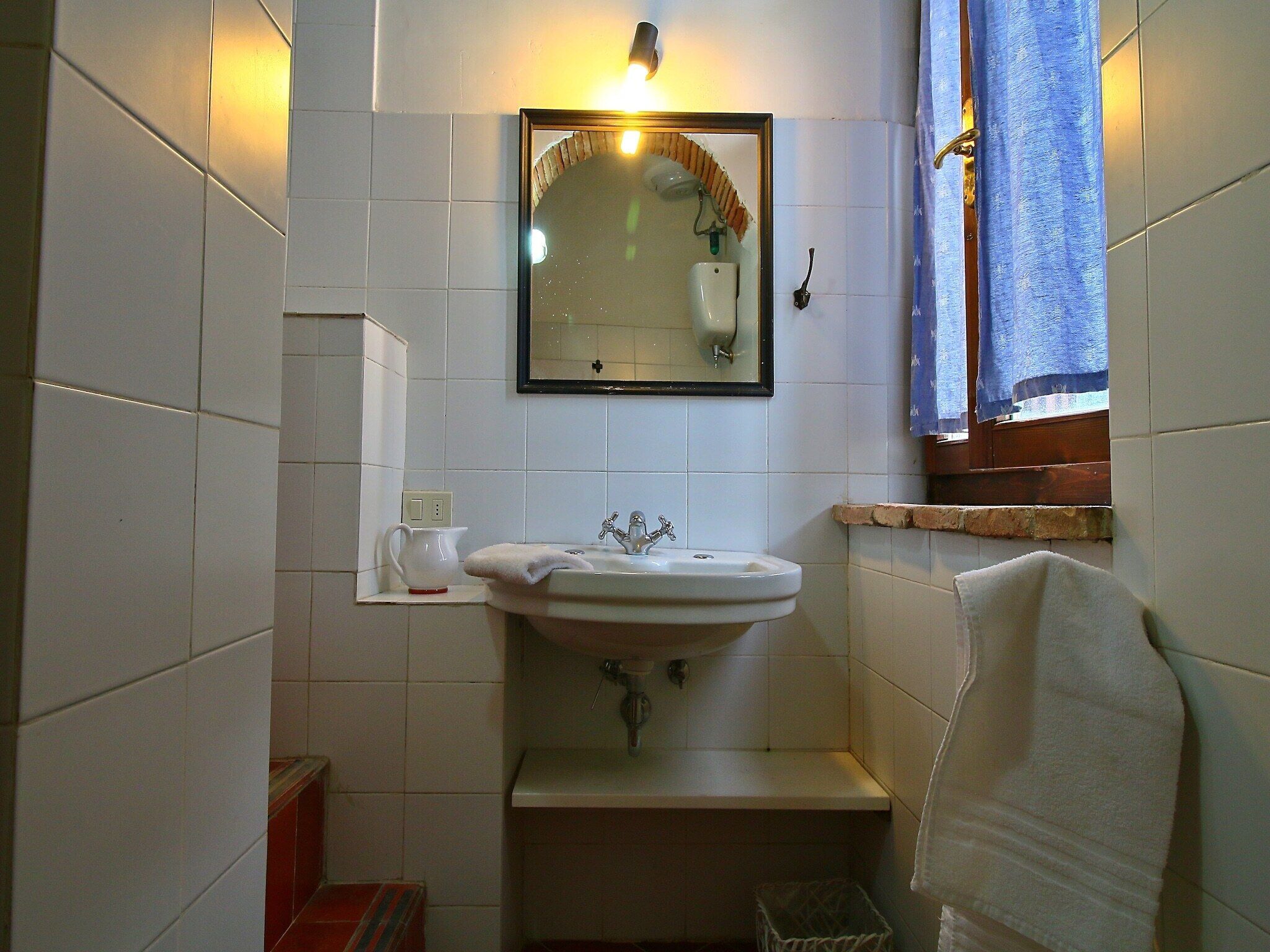 Een kleine badkamer met witte tegels, een wastafel, een spiegel, een handdoek en een raam met blauwe gordijnen.