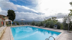 Una piscina rectangular con superficie de agua azul cristalina, rodeada por una solana con baldosas. Hay sillas de descanso y sombrillas a lo largo de la piscina.