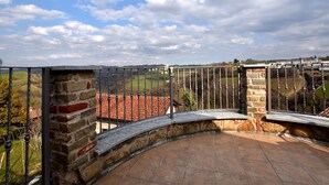 A stone balcony with a metal railing overlooking a rural landscape with fields and a distant building. - Hillview Langhe Apartment (Belvedere Langhe)