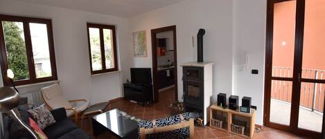 Una sala de estar con un sofá, una mesa de centro y un televisor. Hay una chimenea con un conducto y un balcón visible a través de la puerta.