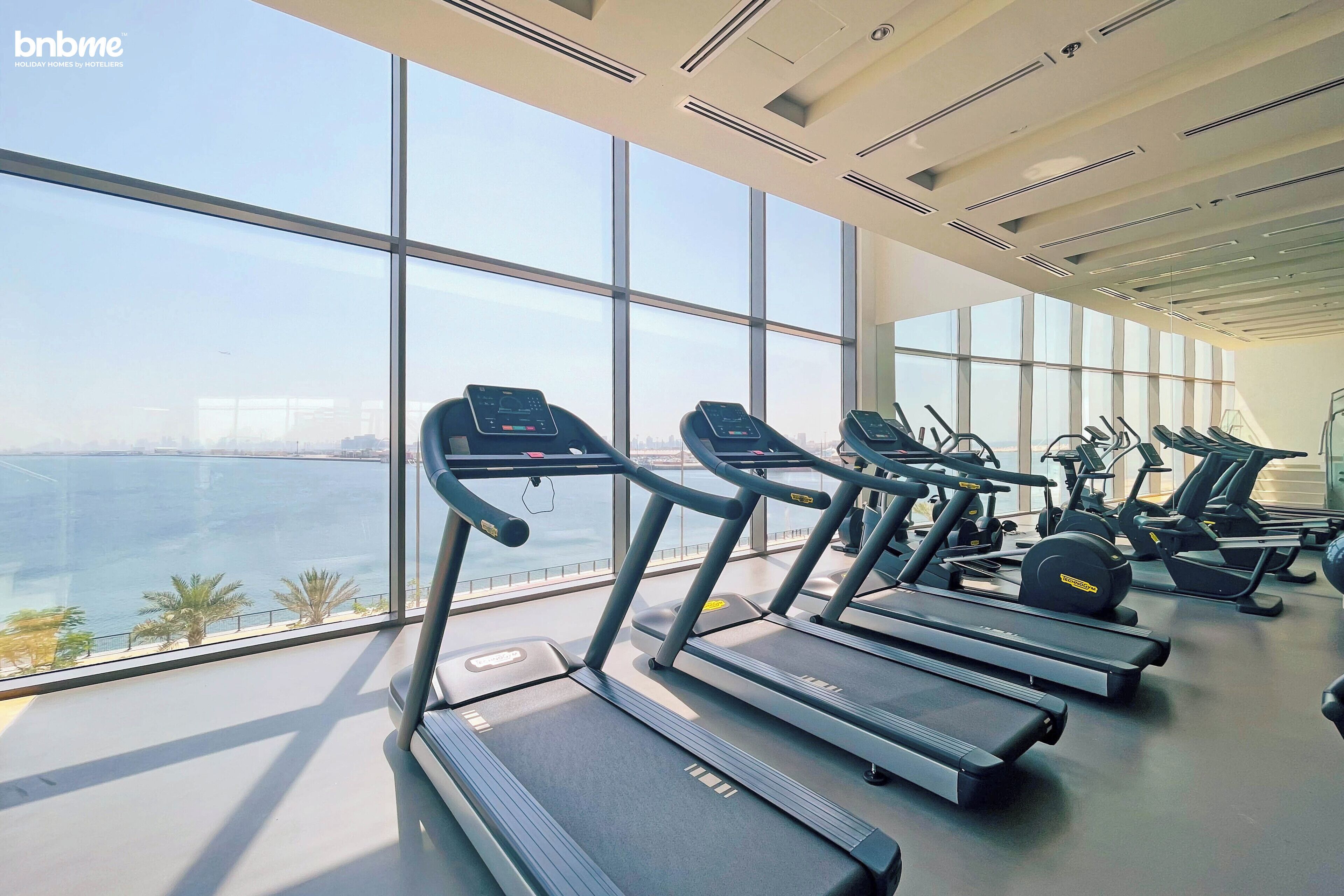 Un gimnasio con cintas de correr y equipo de ejercicio, amplias ventanas con vistas al mar y al paisaje urbano.