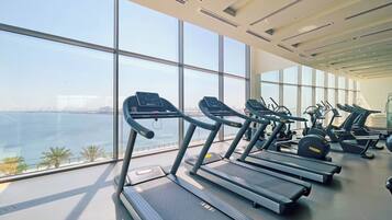 Un gimnasio con cintas de correr y equipo de ejercicio, amplias ventanas con vistas al mar y al paisaje urbano.