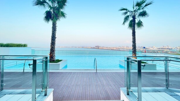 Zona junto a la piscina con barandas de vidrio, palmeras y vistas de la ciudad.