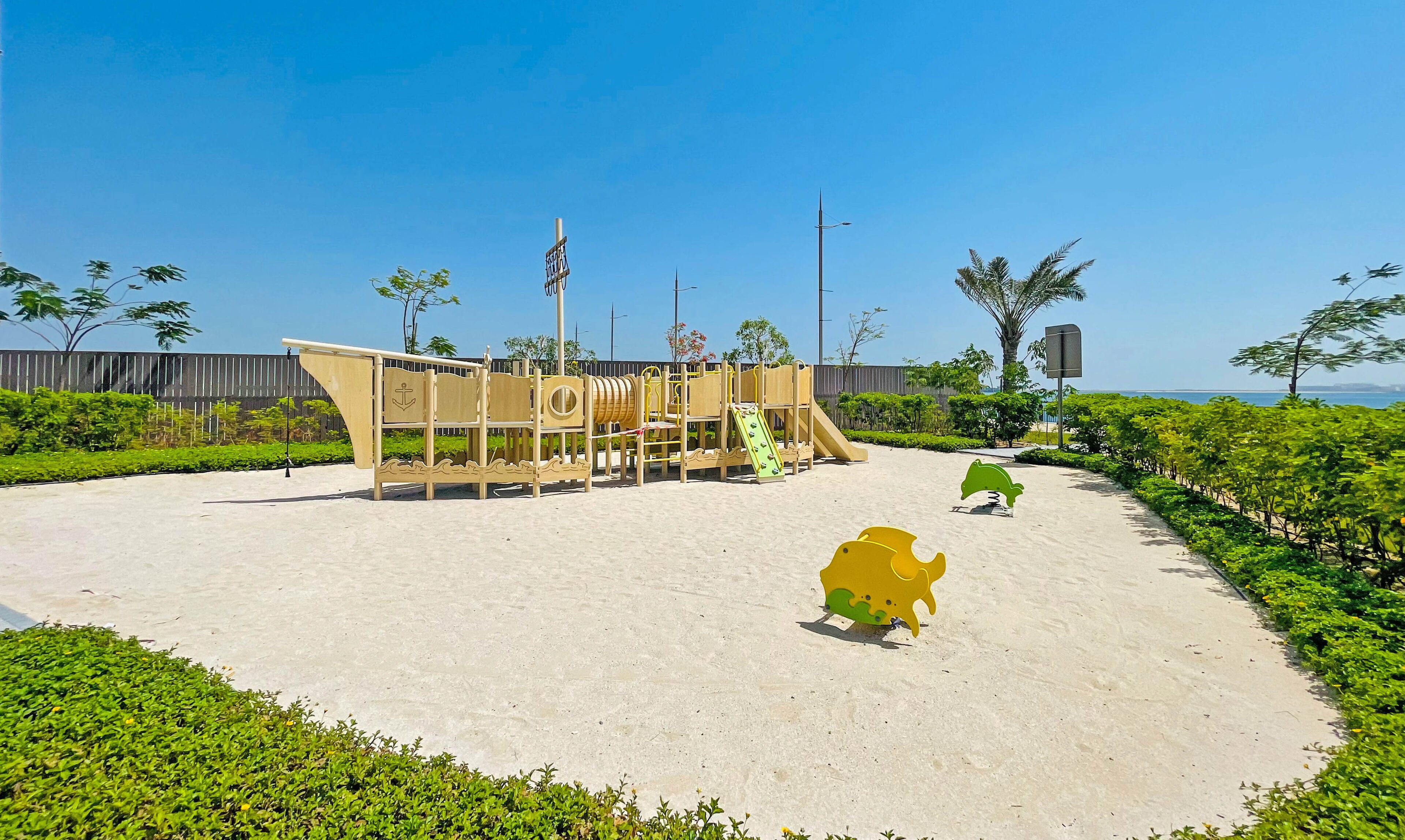 A playground with climbing structures and slides, surrounded by greenery and a clear sky.