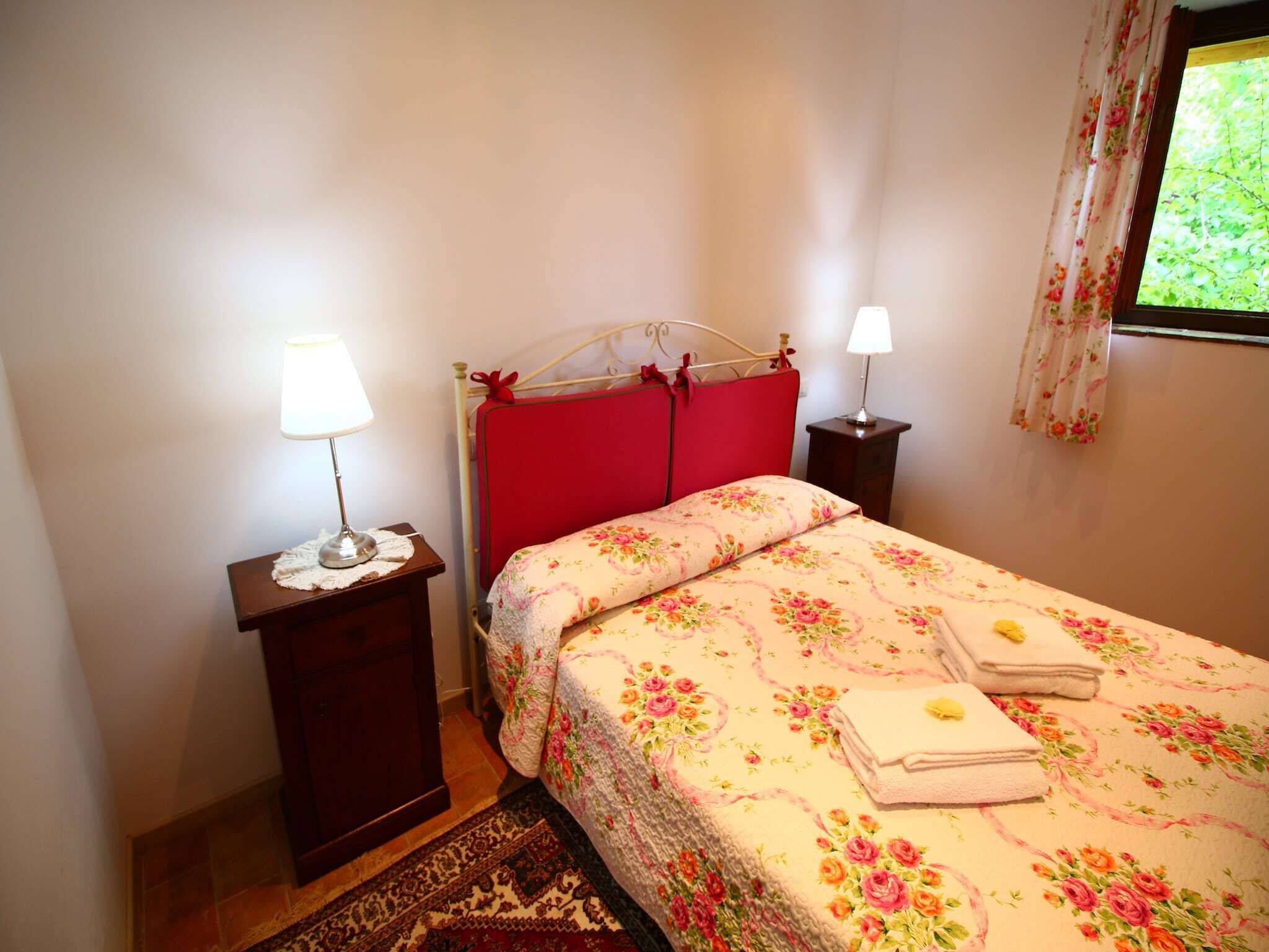 A bedroom with a red headboard, floral bedding, two bedside lamps, and a window with floral curtains.