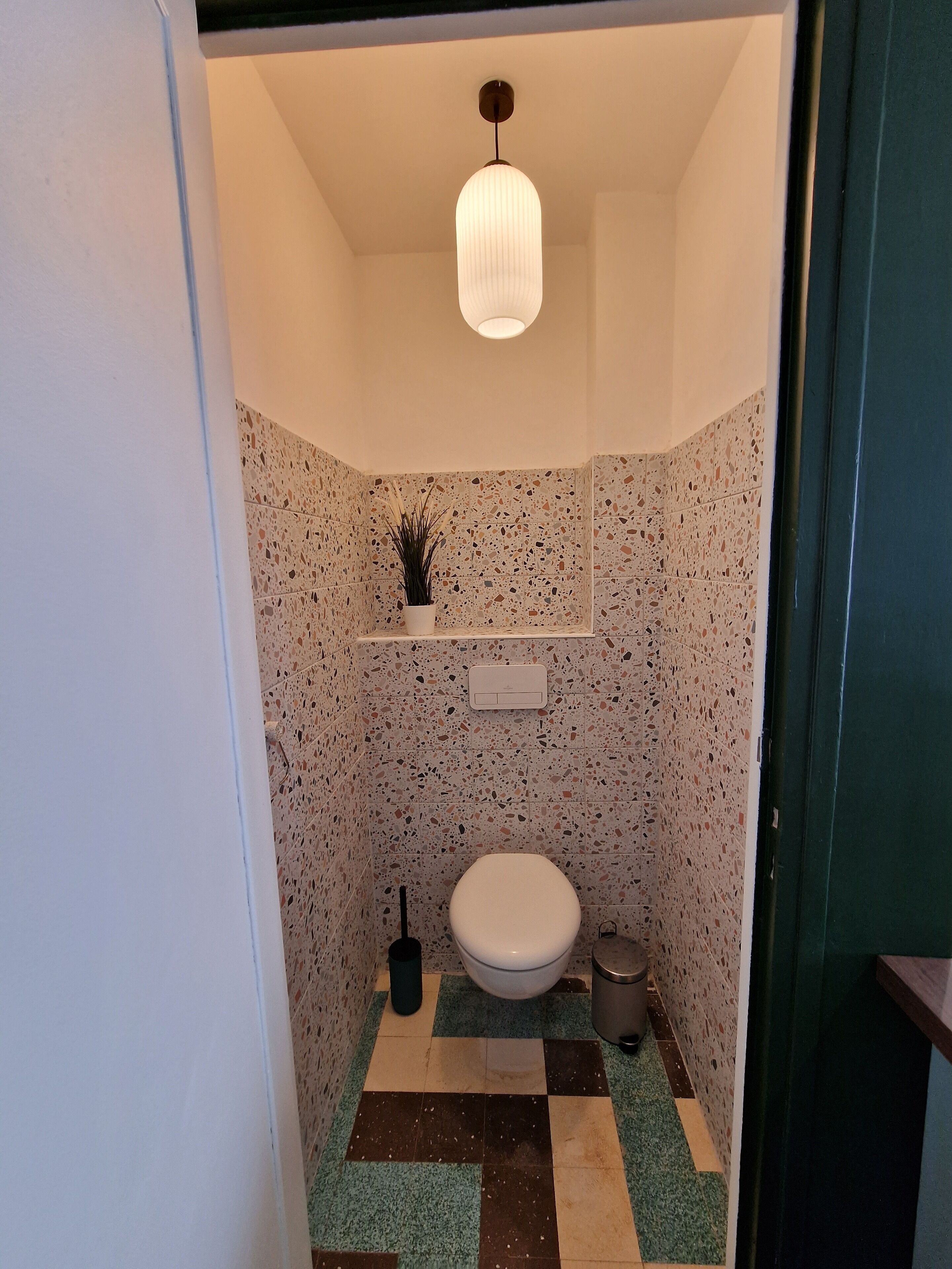 Ein kleines Badezimmer mit Toilette, einem Regal mit Topfpflanze, einer Pendelleuchte und einem Schachbrettfußboden.