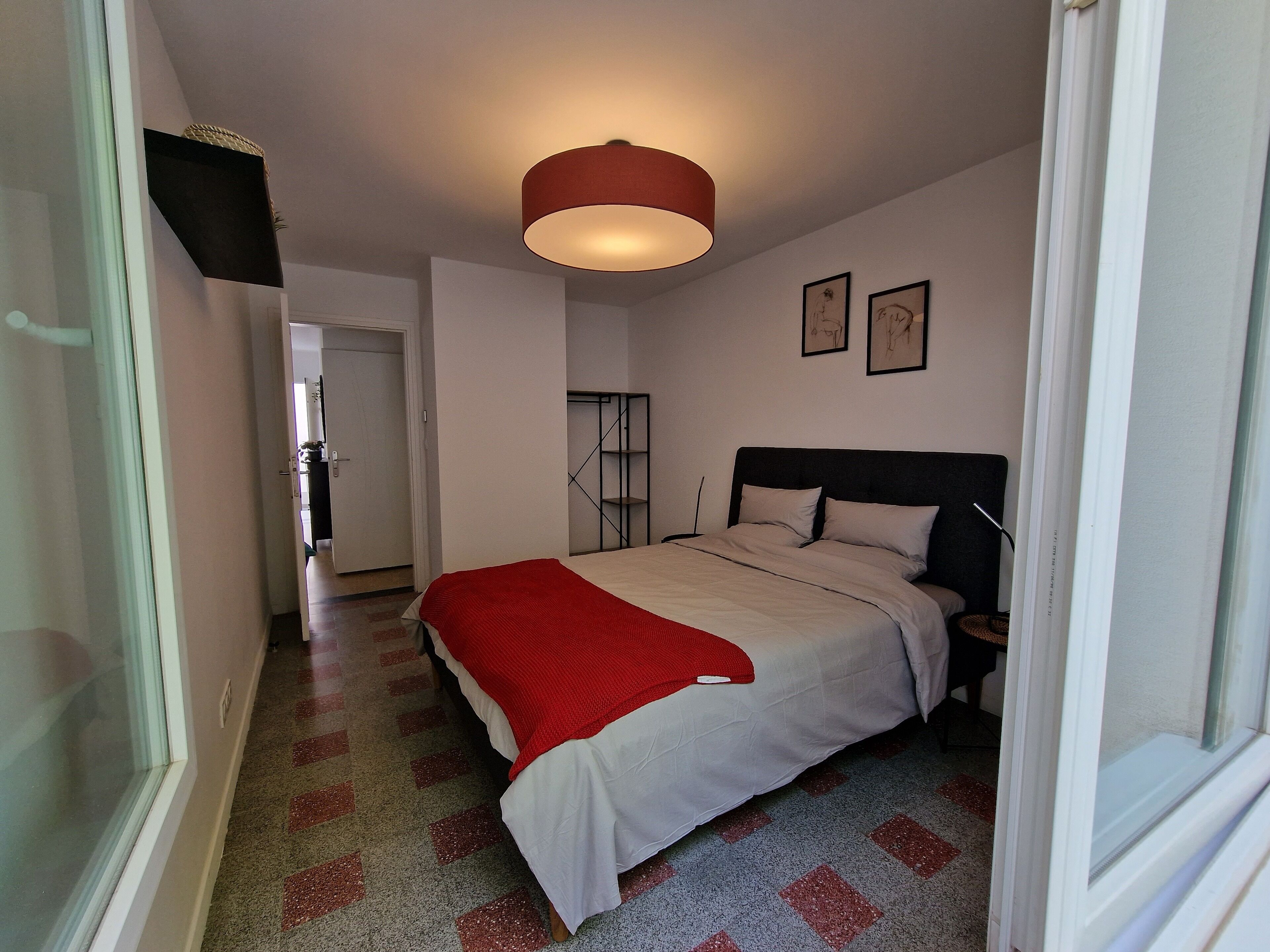 A bedroom with a bed, a bedside table, a red lamp, and a door leading to another room.