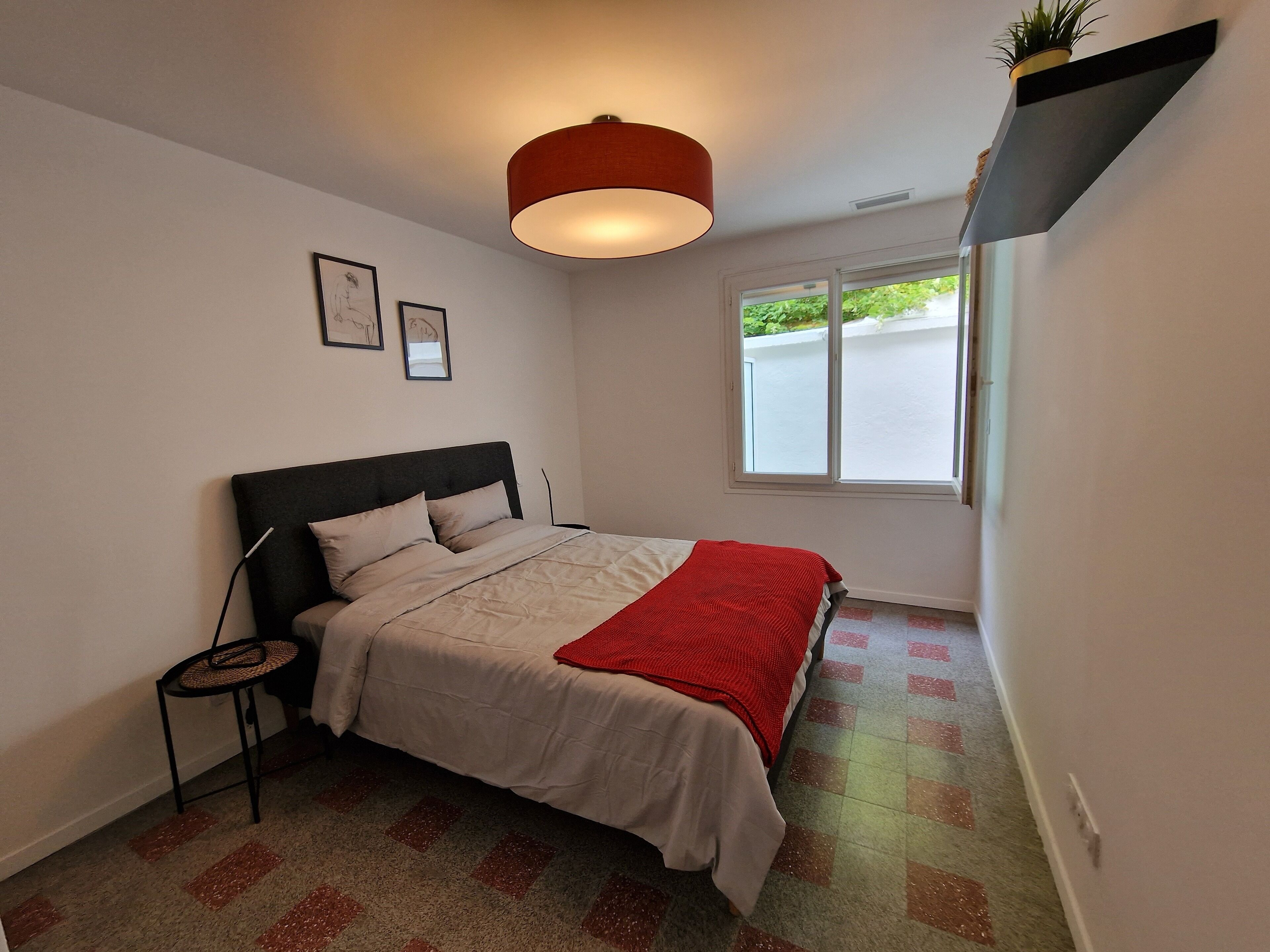 Een slaapkamer met een bed, een nachtkastje, een hangplant, een raam en een rode lamp.