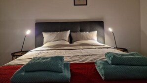 Een netjes opgemaakt bed met witte lakens en twee kussens, een rode deken, en twee opgevouwen handdoeken. Er staan twee bedlampen en twee kunstwerken in lijsten aan de muur.