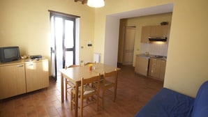 Una cocina compacta con armarios de madera, una mesa de comedor con sillas, un televisor sobre un mueble bajo y un sofá azul en la sala.