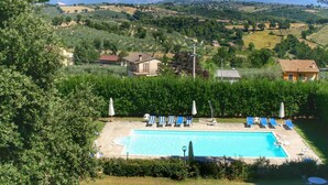 Una piscina con sillas de descanso y sombrillas, rodeada de vegetación y casas, con montañas al fondo.