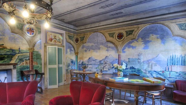 Una habitación con sillones de terciopelo rojo, una mesa de madera con libros, una araña y un mural en la pared.