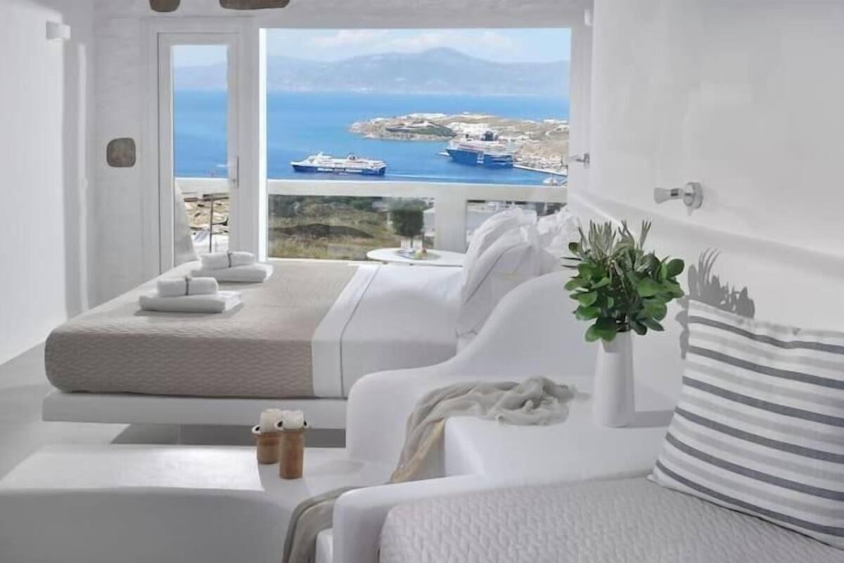 Une chambre moderne et minimaliste, dotée d’une grande fenêtre donnant sur la côte, et comprenant deux lits, une petite table et une plante en pot.