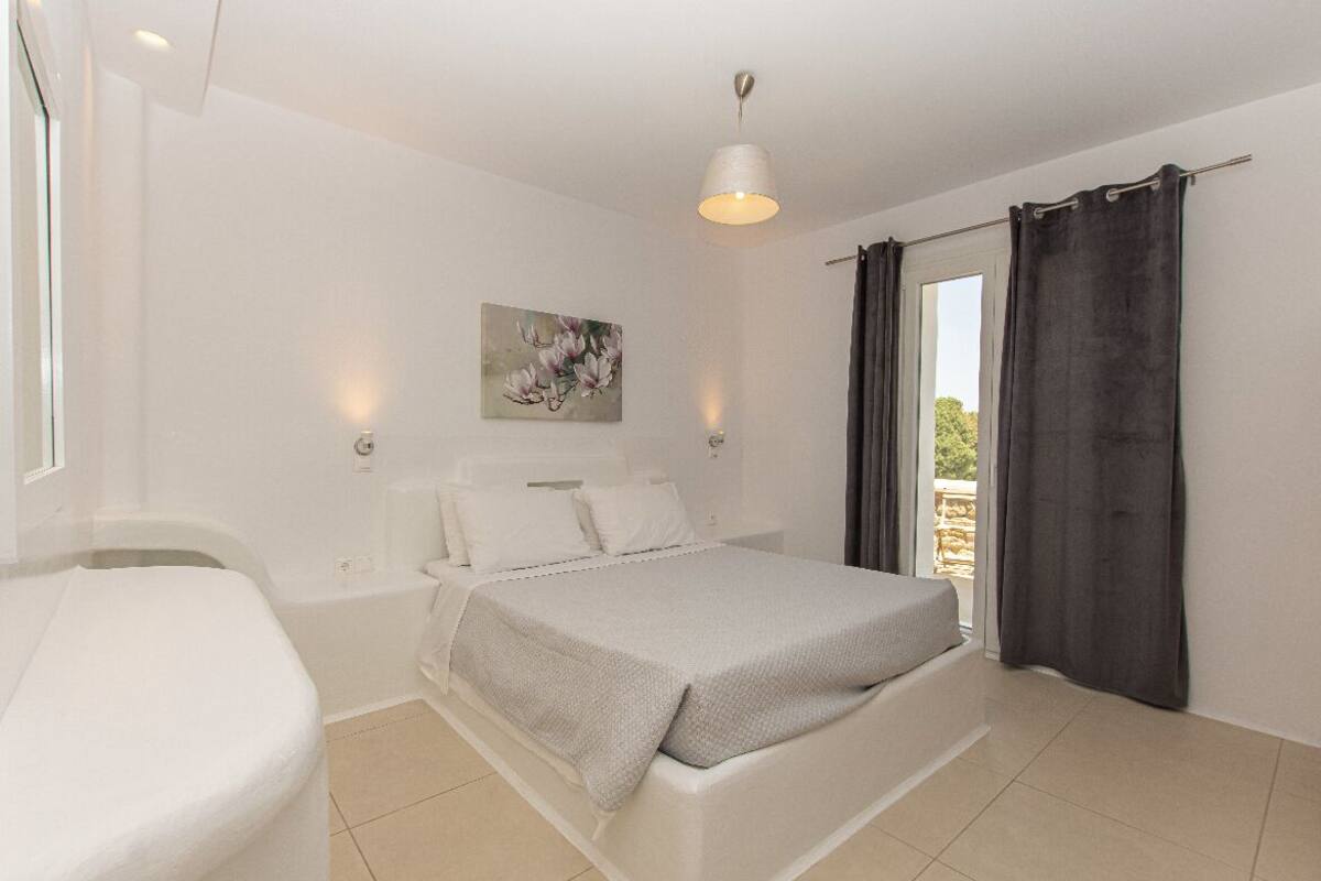 Une chambre à coucher avec un lit, une table de chevet, un tableau, un luminaire au plafond et une vue par une fenêtre avec des rideaux.