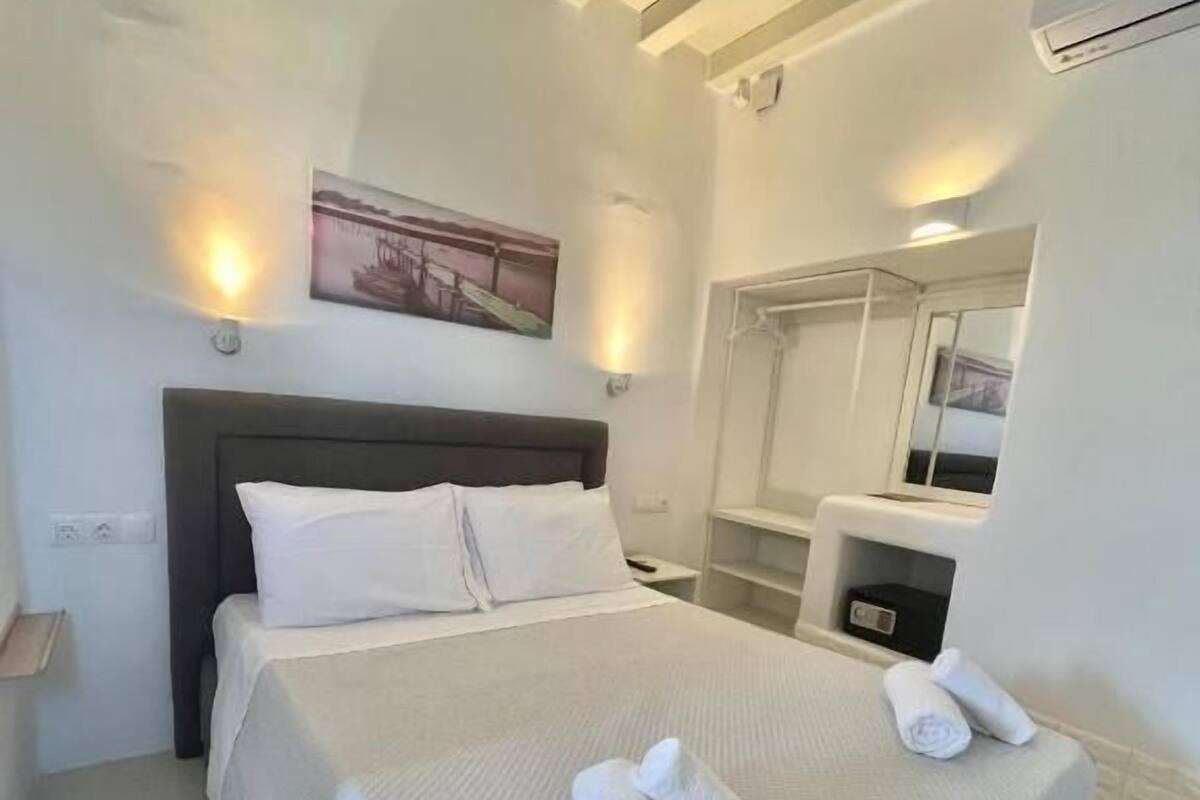 Un lit bien fait, avec du linge de lit et des serviettes blanches, une tête de lit, une table de chevet avec une lampe et un climatiseur fixé au mur.