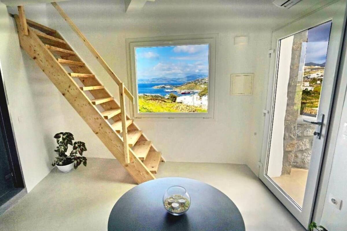 Une pièce avec un escalier en bois, une table ronde avec un plateau en verre, une plante en pot et une fenêtre donnant sur la côte.