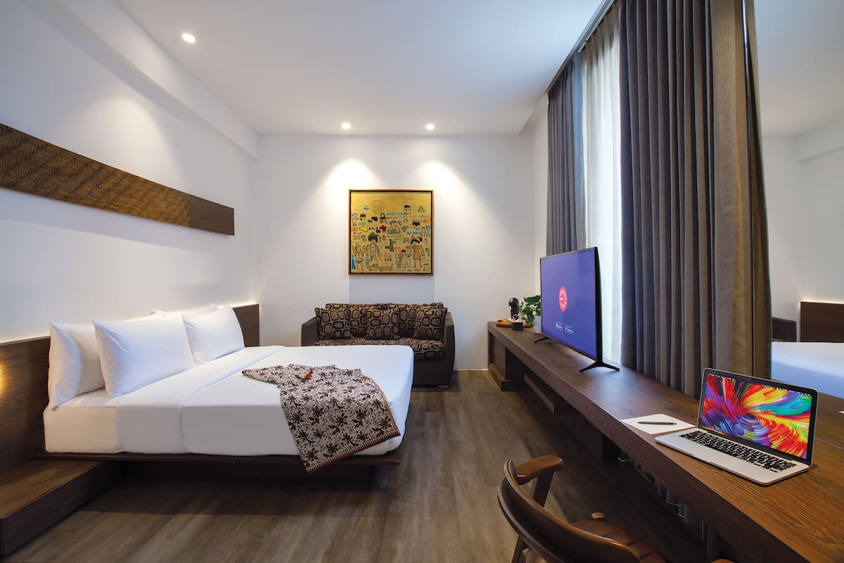 A modern hotel room with a bed, a sofa, a desk with a laptop, a TV, and a large artwork on the wall.