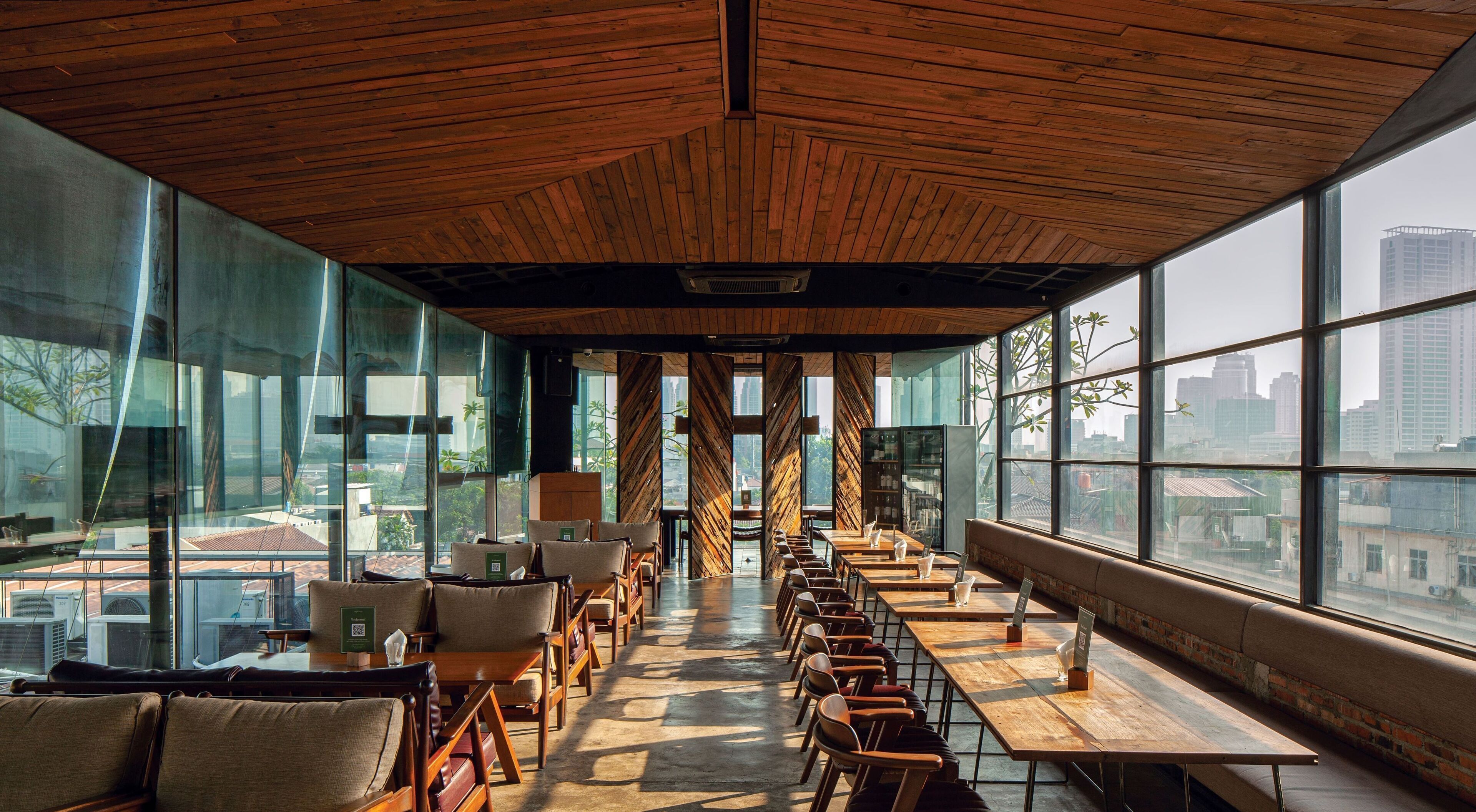 A modern dining area with wooden tables and chairs, large windows offering city views, and a wooden ceiling.
