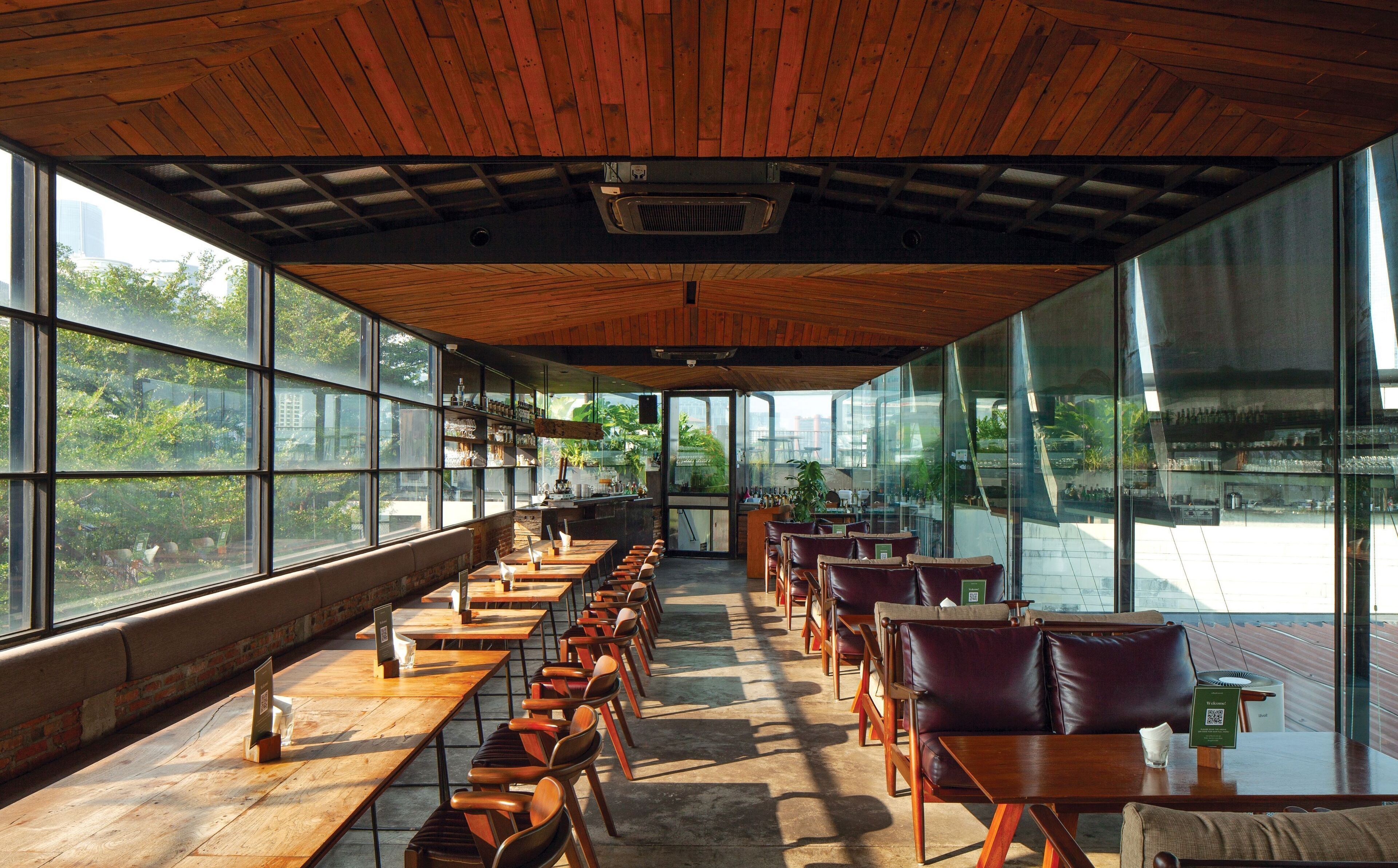 Un vaste espace intérieur aménagé avec de longues tables en bois et des fauteuils en cuir, de grandes fenêtres offrant une vue sur la verdure et les bâtiments, et un plafond en bois doté d’un système de ventilation.