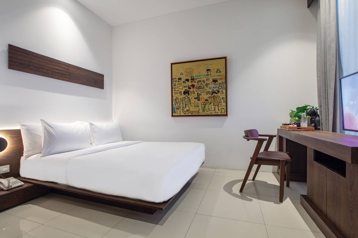 A modern hotel room with a large bed, a desk with a chair, a small plant, and a wall-mounted artwork.