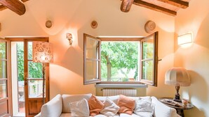 A cozy living room with a beige sofa, a wooden ceiling, and a large window with a view of greenery. - Scenic Stay in Restored Retreat (Arezzo)