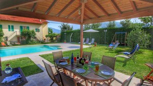 Ein überdachter Essbereich im Freien mit Pool und Schaukeln im Hintergrund.