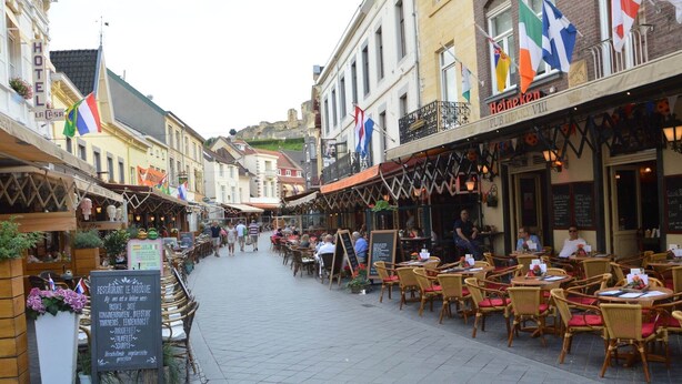 Una strada con tavoli all'aperto nei caffè e ristoranti, un cartello per un ristorante e bandiere sugli edifici.