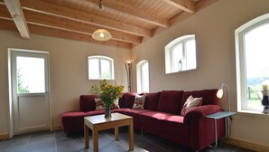 A living room with a sectional sofa, a coffee table, and a vase of flowers. There are windows with white frames and a wooden ceiling. - Holiday Home in Holten near Lake IJssel (Holten)