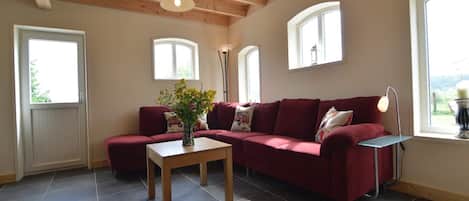 A living room with a sectional sofa, a coffee table, and a vase of flowers. There are windows with white frames and a wooden ceiling.