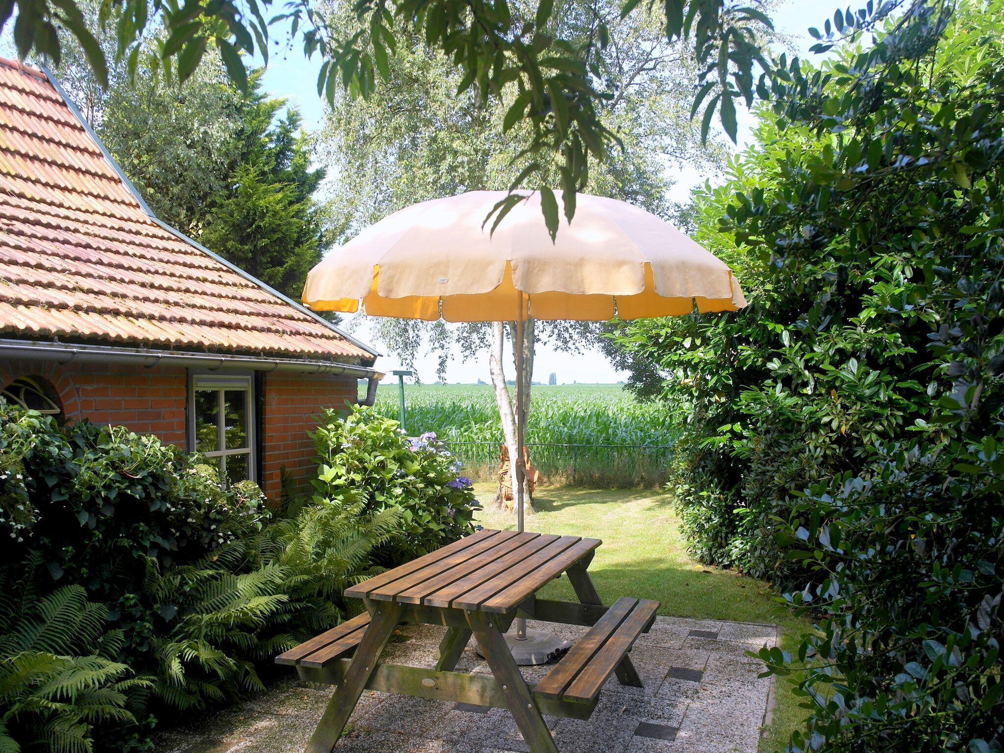 A wooden picnic table with benches, a yellow umbrella, and a brick house with a tiled roof.