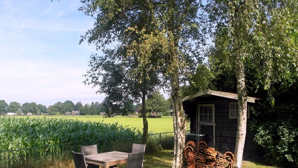 A rustic outdoor seating area with a wooden table and chairs, surrounded by a lush green field and trees.