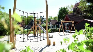 A playground with a netted climbing structure, a slide, and swings, surrounded by sand and greenery. - Detached Home in Netherlands with Garden (Dalfsen)