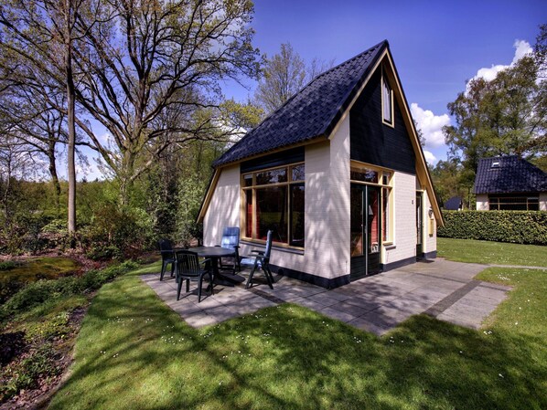 A small, single-story house with a sloped roof, large windows, and a patio with outdoor furniture.