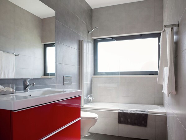 A modern bathroom with a red vanity, a bathtub, and a window providing natural light.