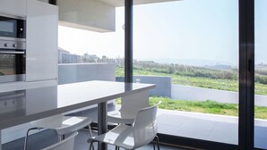 A modern kitchen with white cabinets and stainless steel appliances, a large dining table with white chairs, and a sliding glass door leading to a balcony with a view of greenery and distant hills. - Villa in Portugal with Sea View & Pool (Lourinhã)