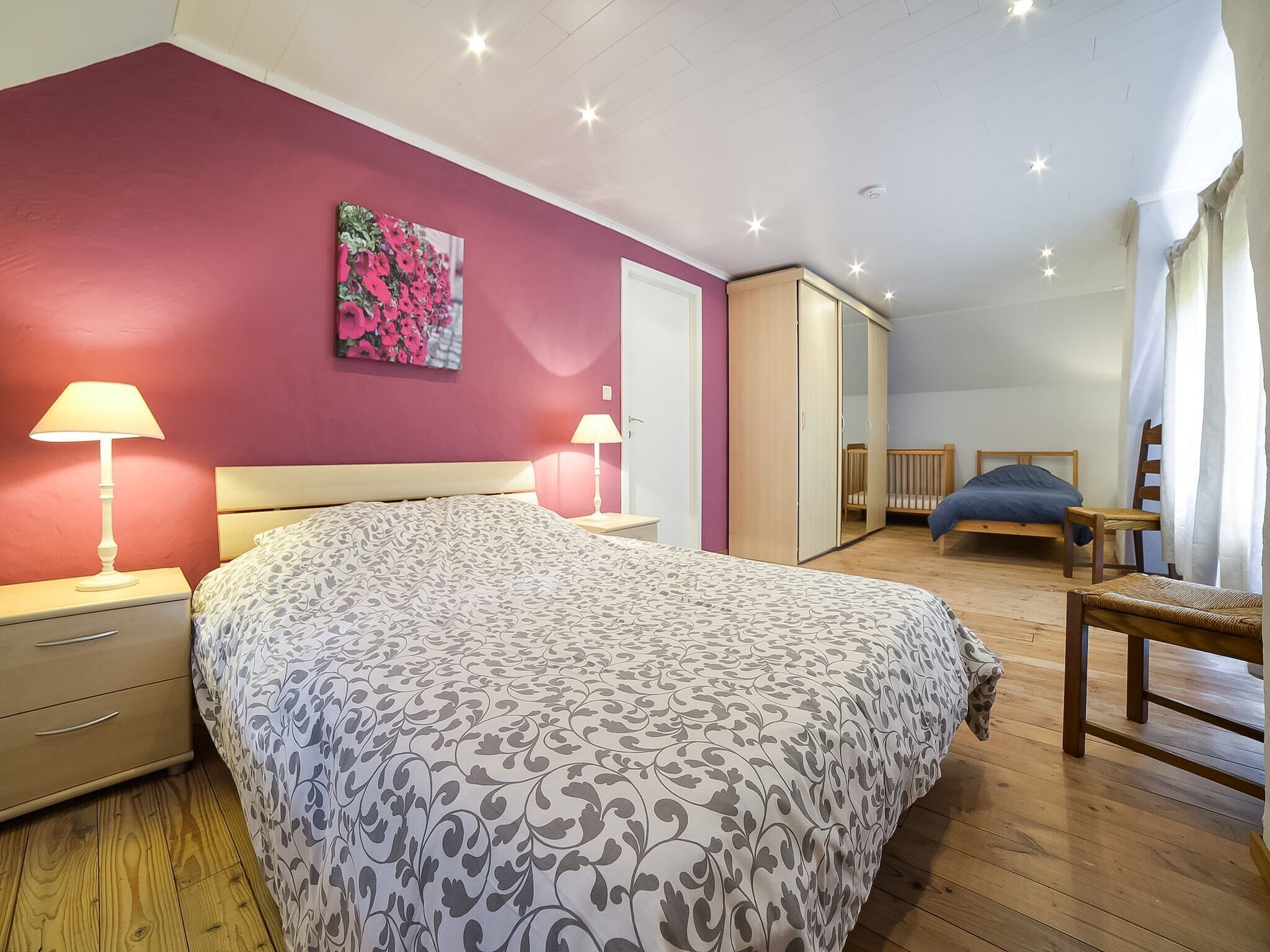 A bedroom with a bed, bedside lamps, a wardrobe, a crib, and a chair.