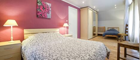 A bedroom with a bed, bedside lamps, a wardrobe, a crib, and a chair.