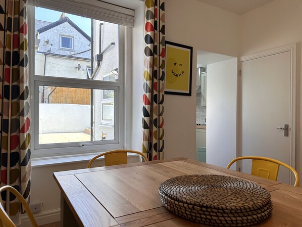 Uma área de refeições com mesa de madeira, cadeiras amarelas, um cesto de vime, uma janela com cortinas e uma cozinha visível através de uma porta aberta.