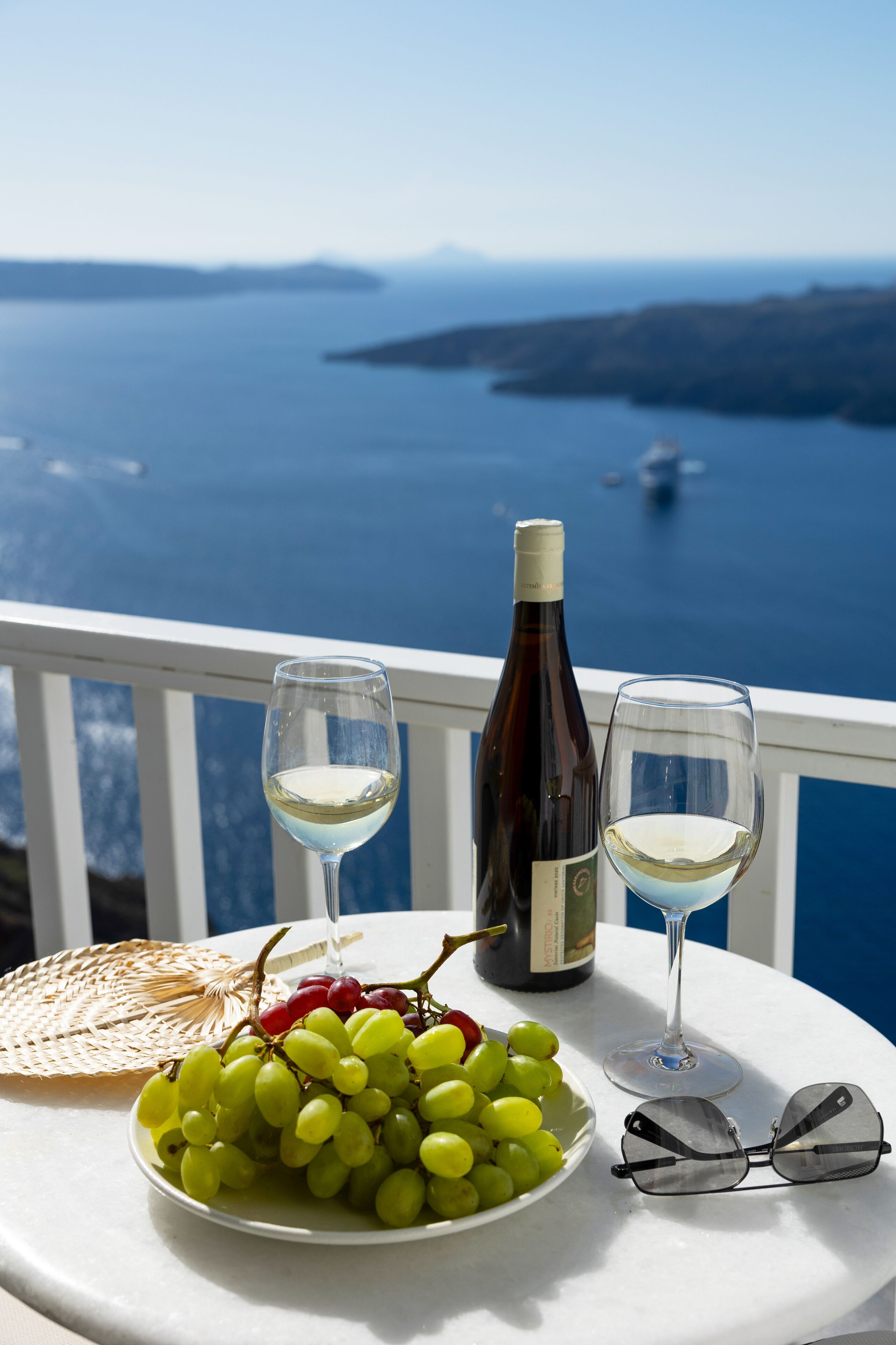 Una mesa con una botella de vino, dos copas de vino, un cuenco de uvas, un plato de pan y un par de gafas de sol en un balcón con vistas al agua.