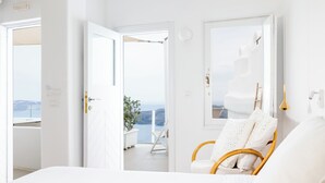 A bright, white room with a bed, a wicker chair, and a view of the sea through an open door.