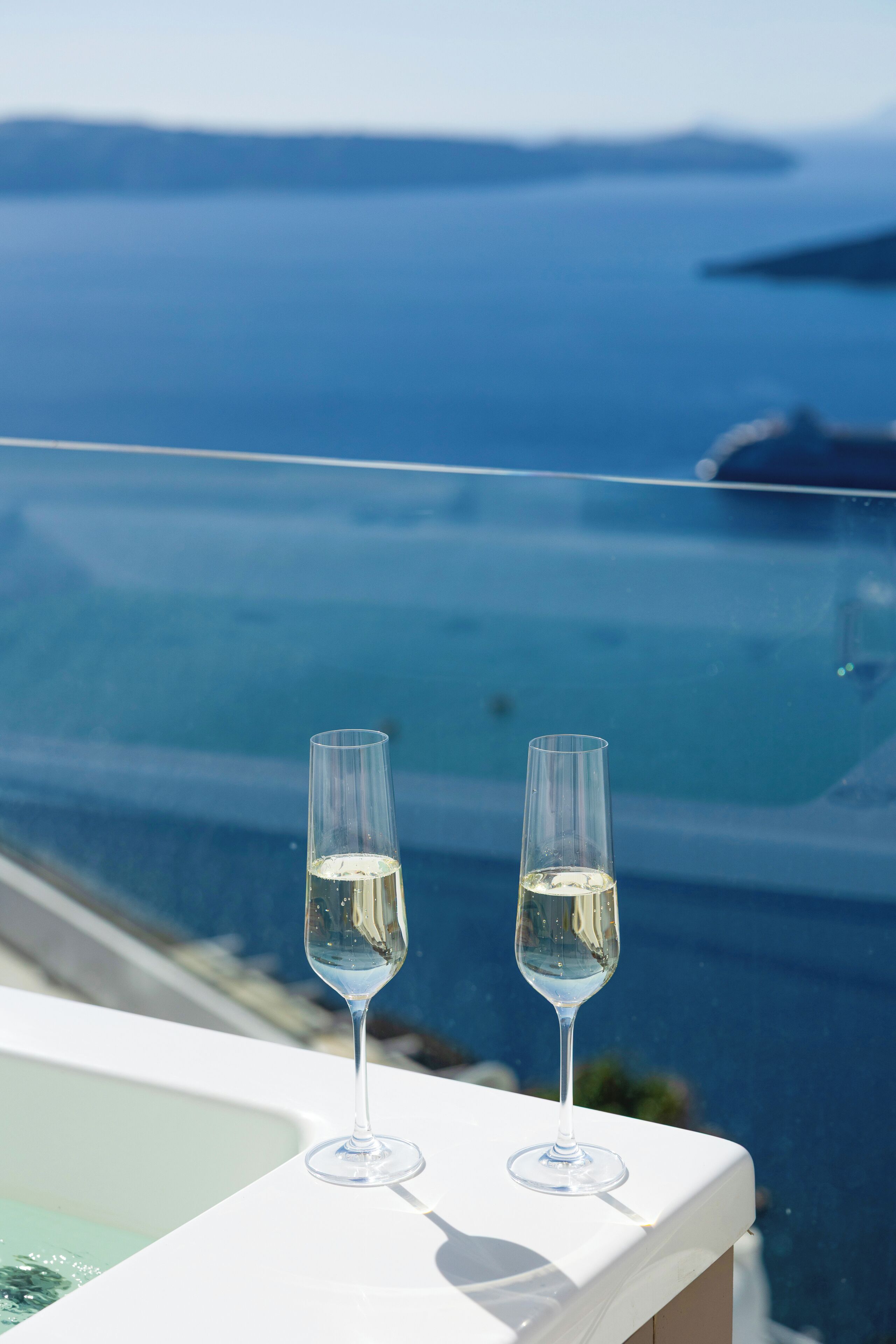Two champagne flutes on a balcony with a view of the sea and distant land.