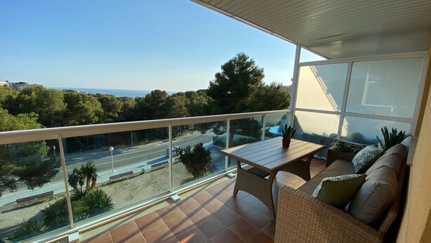 A balcony with a view of a road and trees, a wicker sofa, and a small table. - Tranquilo Apartamento con Vistas al mar y Piscina en Cap Salou (CT)