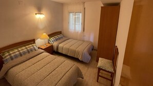 A bedroom with two beds, a nightstand with a lamp, a window with curtains, and a wooden door. - Tranquilo Apartamento con Vistas al mar y Piscina en Cap Salou (CT)