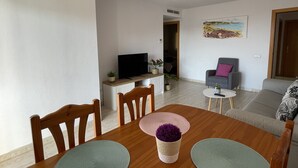 A dining area with a wooden table, chairs, and a TV on a stand. There is a sofa and a painting on the wall. - Tranquilo Apartamento con Vistas al mar y Piscina en Cap Salou (CT)