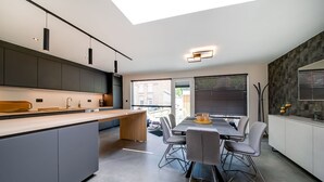 Una cocina moderna con una isla central, una mesa de comedor y sillas. Hay un tragaluz sobre la cocina y una ventana con persianas a la derecha.
