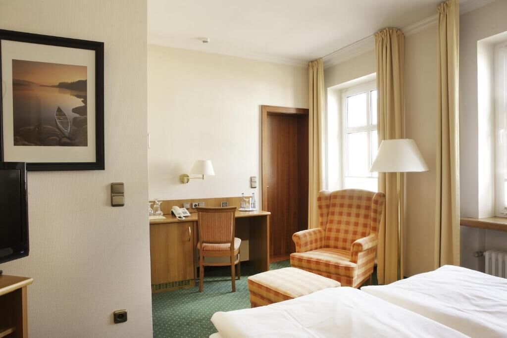 Habitación de hotel con cama, escritorio con silla, televisor, un cuadro en la pared y ventana con cortinas.