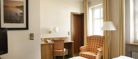 Habitación de hotel con cama, escritorio con silla, televisor, un cuadro en la pared y ventana con cortinas.