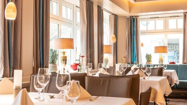 Un restaurant avec de nombreuses tables dressées pour le service, de grandes fenêtres avec des rideaux et un éclairage d’ambiance.