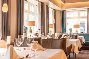 Un restaurante con varias mesas dispuestas para comer, ventanas amplias con cortinas e iluminación ambiental.