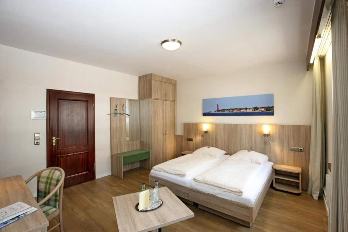 A hotel room with two beds, a desk, a chair, a wooden wardrobe, a small table with a candle, and a painting of a coastal scene.